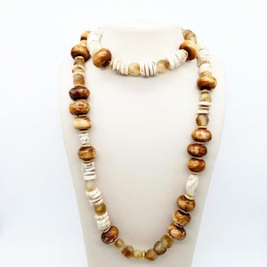 May include: A two-strand necklace displayed on a white mannequin. The necklace features a mix of brown, tan, and white beads in various shapes and sizes. The beads include round, disc, and cylindrical shapes, creating a layered and textured look. The necklace is a statement piece.