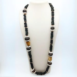 Puede incluir: Un collar largo con cuentas de color verde oscuro y negro, intercaladas con cuentas blancas y marrones estampadas, y piedras de color ámbar. El collar se exhibe en un maniquí blanco, mostrando su longitud y diseño.
