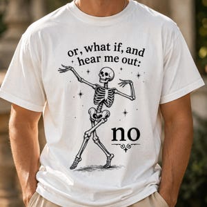 May include: A white short-sleeved t-shirt featuring a black graphic of a dancing skeleton. The text above the skeleton reads, or, what if, and hear me out: and the word no is printed in large black letters below the skeleton with decorative lines.