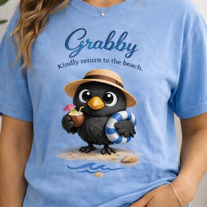 May include: Light blue t-shirt featuring a cartoon bird wearing a straw hat and holding a coconut drink and a blue and white striped inflatable ring. The shirt has the text "Grabby" and "Kindly return to the beach."