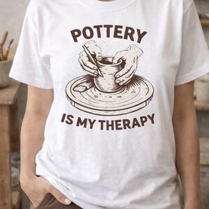 May include: White t-shirt featuring a brown illustration of hands working with clay on a pottery wheel. The text "POTTERY IS MY THERAPY" is printed above and below the graphic in brown. The shirt is short-sleeved.