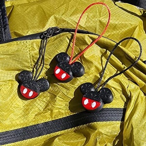 May include: Three black and red Mickey Mouse-shaped zipper pulls with white accents are displayed against a yellow background. Two pulls have red cords, and one has a black cord. The zipper pulls are attached to a yellow bag with black zippers.