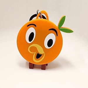 May include: Orange plush keychain featuring a cartoon orange character with large black and white eyes, a yellow beak, and green leaf accents. The keychain has a metal ring and a small brown base.