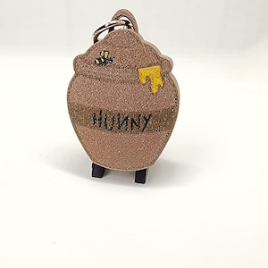 May include: A brown keychain shaped like a honey pot with the word "HUNNY" printed on it. The pot has a yellow honey drip and a small bee graphic. The keychain is on a white background.