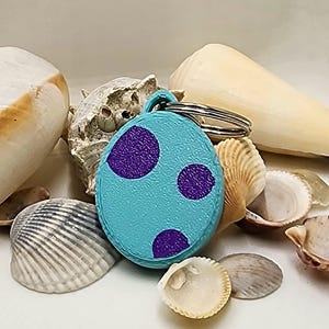 May include: A circular, turquoise keyring with three large purple polka dots. The keyring is made of a textured material and has a silver ring attached. The background features various seashells.