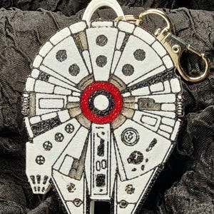 Puede incluir: Un llavero blanco y negro del Halcón Milenario con un círculo central rojo y un cierre dorado. El llavero es una réplica detallada de la icónica nave espacial de la franquicia Star Wars.