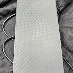 Puede incluir: Un dispositivo rectangular gris con un patrón de tejido de fibra de carbono. El dispositivo tiene esquinas redondeadas y está conectado a un cable negro. El fondo es una tela oscura y texturizada.