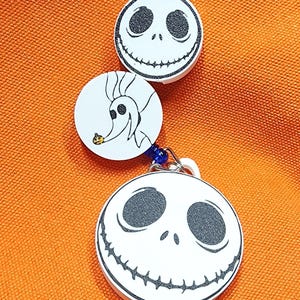 May include: Three white circular charms with black and white character designs from The Nightmare Before Christmas. The top charm features Jack Skellington's face, the middle charm features Zero, and the bottom charm features Jack Skellington's face.