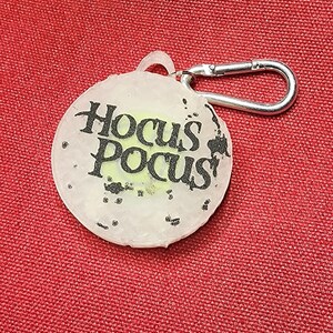 May include: A round, clear keychain with the words "Hocus Pocus" in black lettering. The keychain has a silver clasp and a small loop for attachment. The background is a textured red fabric.