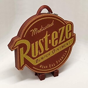May include: A brown and gold Rust-eze sign with a handle. The sign has the words "Medicated Rust-eze Bumper Ointment Rear End Formula" in gold lettering. The sign is on a brown stand.