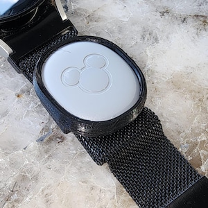 May include: A black watch band with a white Mickey Mouse logo on a round, gray, plastic charm.