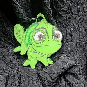 May include: A green chameleon keychain with a cartoon design. The keychain features a bright green body, large brown eyes, and a curled tail. The design has black outlines and a small metal loop for attaching to keys or a bag.