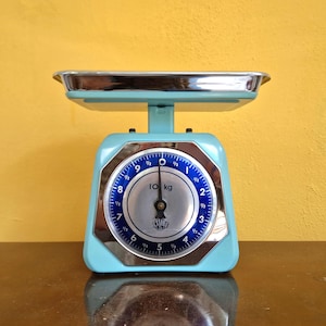 Może przedstawiać: Niebieska waga kuchenna w stylu vintage z chromowaną szalką. Tarcza wagi pokazuje pomiary do 10 kg, z niebieską i srebrną tarczą. Waga stoi na żółtym tle.