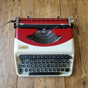 Peut inclure: Machine à écrire vintage rouge et crème. Les touches noires portent des caractères blancs. Le chariot est chromé et le nom "annabella" est visible. Posée sur une surface en bois, cette machine est un outil d'écriture classique.