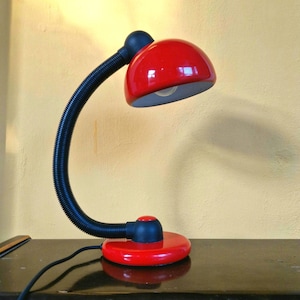 Könnte beinhalten: Eine rot-schwarze Schreibtischlampe mit einem flexiblen, gerippten Hals. Die Lampe hat einen runden roten Sockel und einen roten Lampenschirm. Die Lampe steht auf einer dunklen Oberfläche.