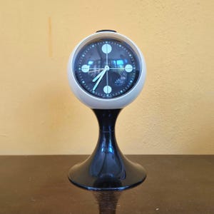 Può includere: Un orologio da tavolo sferico con cassa bianca e base a piedistallo nera. Il quadrante è nero con indicatori circolari bianchi ai punti cardinali, lancette bianche e il testo 'Kienzle' e 'Swiss Made' stampato sul quadrante.