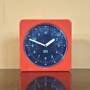 Könnte beinhalten: Eine quadratische orangefarbene Analoguhr mit schwarzem, rundem Zifferblatt. Die Uhr hat weiße Stundenzahlen von 1 bis 12, weiße Minutenmarkierungen von 5 bis 60 und weiße Zeiger. Der Schriftzug "BOSCH electronic" ist in Weiß mittig aufgedruckt.