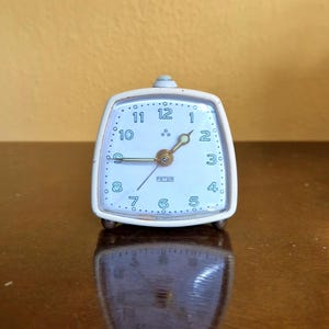 Könnte beinhalten: Ein weißer Vintage-Wecker mit quadratischem Zifferblatt und abgerundeten Ecken. Das Zifferblatt ist hellblau mit schwarzen Ziffern und Zeigern. Das Wort "PETER" ist auf dem Zifferblatt aufgedruckt. Die Uhr hat einen kleinen Knopf oben und steht auf zwei kleinen Füßen.