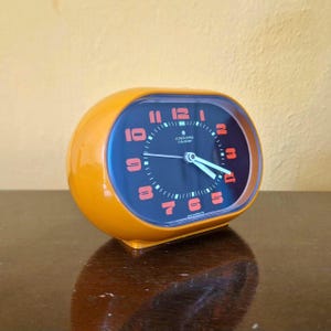 Könnte beinhalten: Orangener Retro-Wecker mit dunkelblauem Zifferblatt und roten Ziffern. Die Uhr hat eine geschwungene, ovale Form und trägt den Markennamen "Junghans Cavalier". Die Zeiger der Uhr sind hellblau.