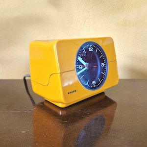 Könnte beinhalten: Ein gelber Vintage-Krups-Wecker mit schwarzem Zifferblatt. Das Zifferblatt hat weiße Zahlen und Zeiger mit einem roten Sekundenzeiger. Die Uhr steht auf einer dunkelbraunen Oberfläche.
