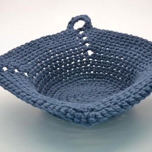 May include: A handcrafted, square crochet basket in a muted blue hue. The basket has a looped handle and a textured stitch pattern. The open weave design provides a visually appealing and practical storage solution.