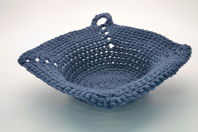 May include: A handcrafted, square crochet basket in a muted blue hue. The basket has a looped handle and a textured stitch pattern. The open weave design provides a visually appealing and practical storage solution.
