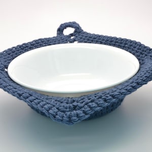 May include: A white ceramic bowl is nestled in a handmade, blue crocheted bowl cosy. The cosy has a square shape with a loop for hanging. The cosy is made of a thick, textured yarn.
