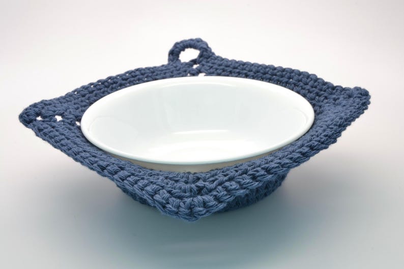 May include: A white ceramic bowl is nestled in a handmade, blue crocheted bowl cosy. The cosy has a square shape with a loop for hanging. The cosy is made of a thick, textured yarn.