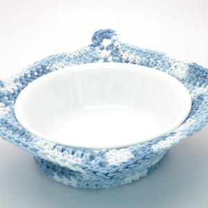 May include: A white ceramic bowl is nestled in a handmade crochet bowl cosy. The cosy is square with a hanging loop, made from blue and white yarn. The bowl is centrally positioned within the cosy.