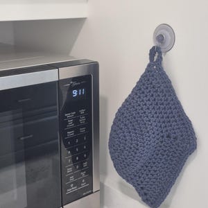 May include: A handmade, blue-grey crochet potholder hanging on a white wall next to a microwave. The potholder is diamond-shaped with a loop for hanging. The microwave displays the time as 9:11.