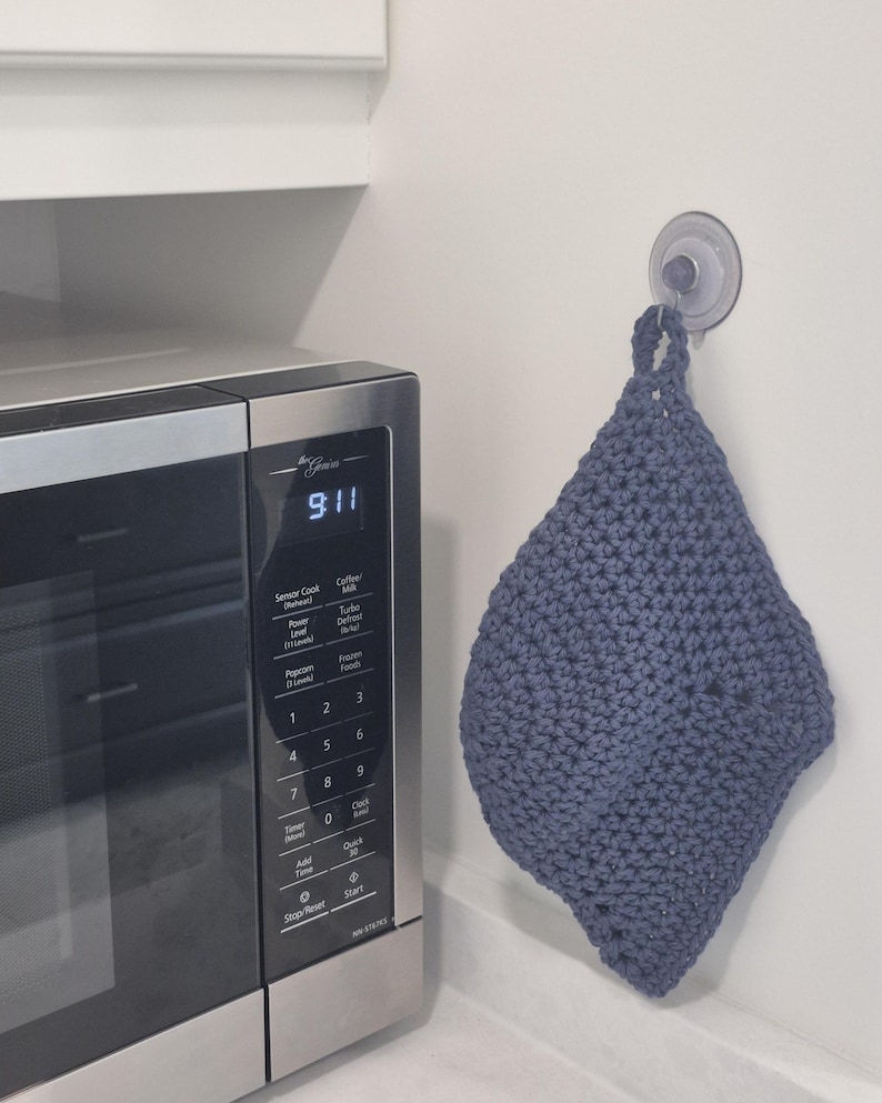 May include: A handmade, blue-grey crochet potholder hanging on a white wall next to a microwave. The potholder is diamond-shaped with a loop for hanging. The microwave displays the time as 9:11.