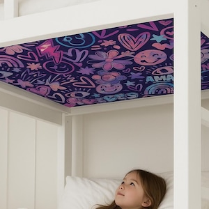May include: A white bunk bed with a colorful, patterned canopy featuring hearts, rainbows, and smiley faces. A young person is lying in the lower bunk, looking up at the canopy. The canopy has the word "AMA" on it.