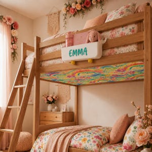 Op de afbeelding: Een houten stapelbed met bloemenbeddengoed en een kleurrijk regenboogpatroon plafond. Een witte opbergcontainer met de naam "EMMA" in turquoise letters is aan het bed bevestigd. Een pluchen konijn en andere items zitten erin.