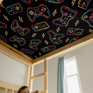 Könnte beinhalten: Ein Kind blickt auf die Decke eines Etagenbettes, die mit einem schwarzen Muster aus farbenfrohen Videospiel-Controllern und Blitzen verziert ist. Die Controller sind rot, gelb und blau umrandet.
