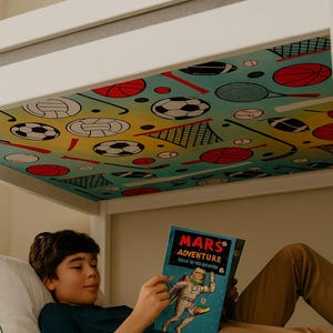 May include: A child reading a book titled "Mars Adventure" under a bunk bed. The bed's underside features a colorful sports-themed pattern with basketballs, soccer balls, and tennis rackets. The child is wearing a blue shirt and brown pants.