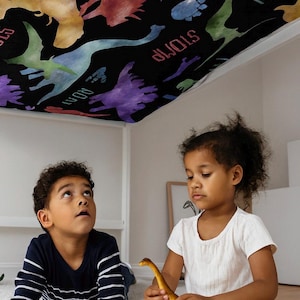 May include: A black dinosaur-themed blanket with colorful dinosaur silhouettes and the words "ROAR", "STOMP", and "RUN". Two children are playing with toy dinosaurs on a white rug. One child is looking up at the blanket.