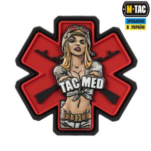 Può includere: Una toppa rossa e nera con un'illustrazione stilizzata di una donna con un casco da pilota, con la scritta "TAC MED". La toppa ha la forma di una croce rossa con due fucili dietro la donna. Il logo M-TAC si trova in alto a destra.