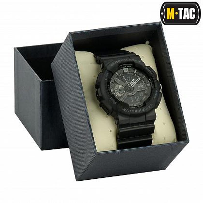 May include: A black digital watch with a black band is displayed inside a gray gift box. The watch face shows the time and has multiple dials and buttons. The box is open, revealing the watch resting on a cream-colored cushion. The watch is water resistant.