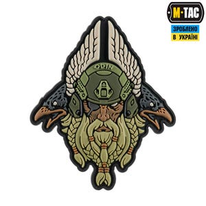 Puede incluir: Un parche que representa a Odín, el dios nórdico, con un casco militar con la palabra "ODIN" y flanqueado por dos cuervos. El parche tiene un borde negro y un diseño detallado con varios tonos de verde, beige y marrón.