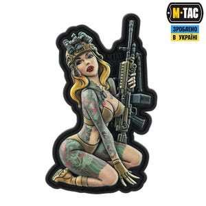 May include: A patch depicting a pin-up style illustration of a woman in military attire, holding a rifle. The woman has blonde hair, red lipstick, and tattoos. The patch has a black border and the M-TAC logo.