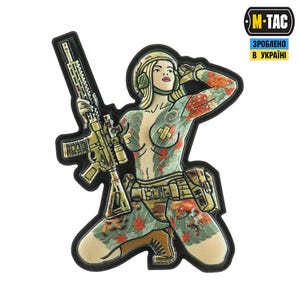 May include: A patch depicting a kneeling woman with tattoos, holding a rifle. The woman wears a helmet and tactical gear. The patch has a black border and the M-TAC logo.