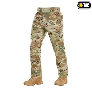 May include: Camouflage pattern pants with multiple pockets and a button closure. The pants are in shades of green, brown, and tan, and are paired with tan-colored boots. The M-TAC logo is visible in the upper right corner.
