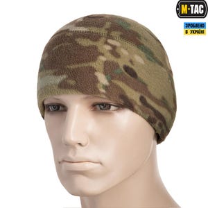 May include: A camouflage pattern beanie hat. The hat is made of a soft, textured material and features a mix of brown, green, and tan colors. The hat is designed to fit snugly on the head. The M-TAC logo is visible in the upper right corner.