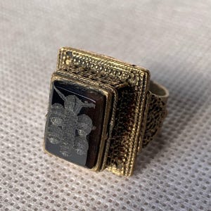 May include: A gold-toned ring with a rectangular face featuring a dark, possibly onyx, stone. The stone is etched with a detailed, stylized image. The ring has a textured band and a decorative setting around the stone.