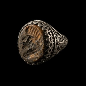 May include: A silver-toned ring featuring an oval-shaped, textured brown stone centrepiece. The metal band displays intricate, ornate filigree patterns along the sides and a scalloped edge surrounding the central stone, creating a detailed appearance.