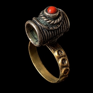 May include: An antique ring with a silver-coloured top and a gold-coloured band. The top features a red stone and intricate detailing. The band has a series of circular designs. The ring is set against a black background.
