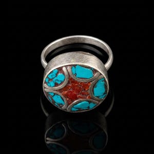 May include: A silver ring featuring a circular face with inlaid turquoise and red stone fragments. The metal has a brushed finish, and the inlay is divided by curved silver lines, creating a geometric pattern within the round setting.