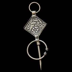 May include: A silver-toned metal brooch featuring a diamond-shaped plaque with intricate floral and leaf engravings. The piece includes a circular ring at the top and a larger circular pin mechanism at the bottom with a pointed needle-like component.