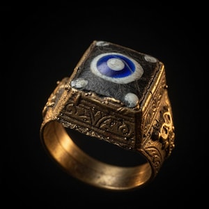 Puede incluir: Un anillo dorado con un diseño ornamentado. El anillo presenta una parte superior rectangular con un fondo negro y un diseño en forma de ojo azul y blanco. La banda y los lados del anillo están detallados con patrones intrincados.