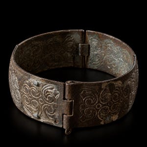 Puede incluir: Una pulsera articulada de metal marrón oscuro con un patrón de volutas en relieve que se repite por toda la superficie exterior. El brazalete está formado por dos segmentos curvos unidos por una bisagra visible con una pequeña anilla en la base.
